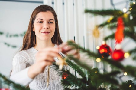 Photo pour Belle souriante femme caucasienne gaie vêtue d'une tenue blanche décorant un arbre de Noël naturel, mettant des ornements rouges sur l'arbre, tenant un ornement de Noël rouge. - image libre de droit