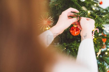 Photo pour Femme caucasienne méconnaissable décorer l'arbre de Noël, mettre ornement rouge sur l'arbre, tenant un ornement de Noël rouge. - image libre de droit