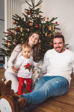 Foto de Feliz joven alegre familia caucásica de tres padres y una niña divirtiéndose decorando el árbol de Navidad. Retrato de Navidad familiar. - Imagen libre de derechos