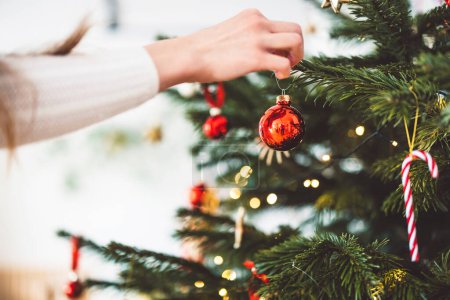 Foto de Mujer caucásica irreconocible decorando el árbol de Navidad, poniendo adorno rojo en el árbol, sosteniendo un adorno rojo de Navidad. - Imagen libre de derechos