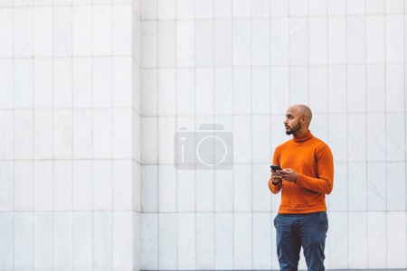 Photo for Caucasian man in an orange sweater standing outside in an urban setting holding a digital device, office buildings in the background. - Royalty Free Image
