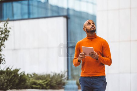 Photo for Caucasian man in an orange sweater standing outside in an urban setting holding a digital device, office buildings in the background. - Royalty Free Image
