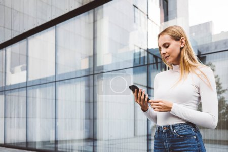 Photo for Young caucasian woman in an urban modern setting, office buildings in the background, using a mobile device. Business woman on a break. - Royalty Free Image