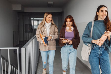Téléchargez les photos : Groupe de trois amies, de jeunes étudiantes caucasiennes marchant à l'intérieur de l'école pendant une pause, se sont dirigées vers une autre classe. Étudiants portant leurs livres avec eux. - en image libre de droit