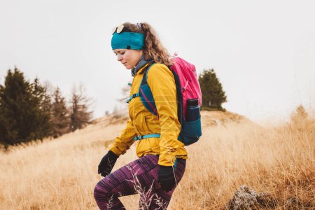 Foto de Caminante mujer caucásica en chaqueta amarilla y mochila rosa senderismo en las montañas en un día de otoño, con guantes y diadema. - Imagen libre de derechos
