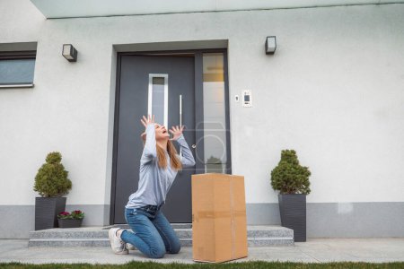 Téléchargez les photos : Thrilled caucasian woman standing at the front door with a big cardboard box that just came in the mail. Woman receiving an exciting package, holding it in her arms. - en image libre de droit