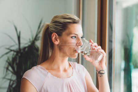 Photo for Smiling young caucasian woman taking her morning supplements, vitamin pills with a glass of water at home. High quality photo - Royalty Free Image