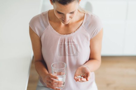 Téléchargez les photos : Souriante jeune femme caucasienne prenant ses suppléments matinaux, des pilules vitaminées avec un verre d'eau à la maison. Photo de haute qualité - en image libre de droit