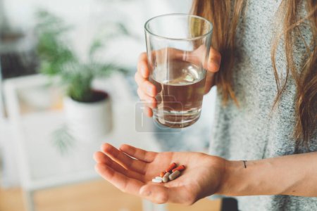 Photo for Smiling young caucasian woman taking her morning supplements, vitamin pills with a glass of water at home. High quality photo - Royalty Free Image