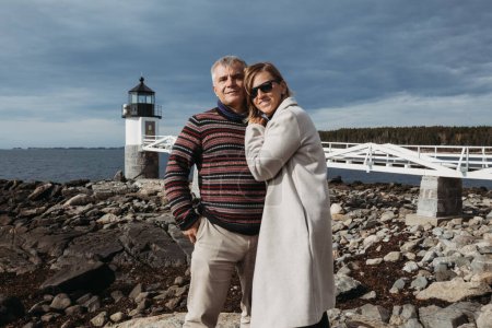 Photo for Middle age couple standing and hugging with lighthouse in the background - Royalty Free Image