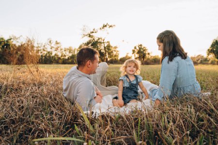 Téléchargez les photos : Jeune couple avec jolie fille souriante de deux ans assise sur l'herbe en tenue bleu clair - en image libre de droit