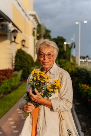 Téléchargez les photos : Femmes âgées tenant des fleurs jaunes et souriant en robe orange - en image libre de droit