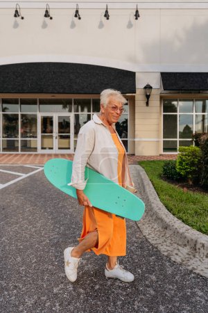 Photo for Elderly women in orange dress and ivory shirt holding skateboard - Royalty Free Image