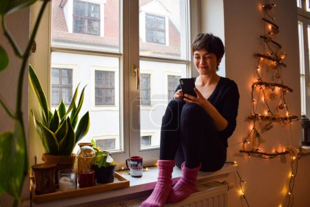 Photo for Young woman makes purchases in the Internet and pays by smartphone on Black Friday. Girl is enjoying buying gifts and sitting on comfortable windowsill at home - Royalty Free Image