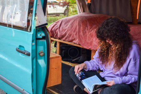 Photo for Independent curly hair backpacker woman laughing laying in bed inside her motorhome while reading a book during an adventure road trip with her camper van listening to music - Royalty Free Image