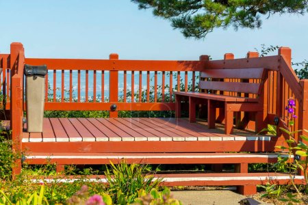 Red wooden and timber balcony with bench and hand rails with sky lined overlook midday in sun with foreground plants and tree. Steps and trash can in public area with deck for standing or smoking in suburban area.