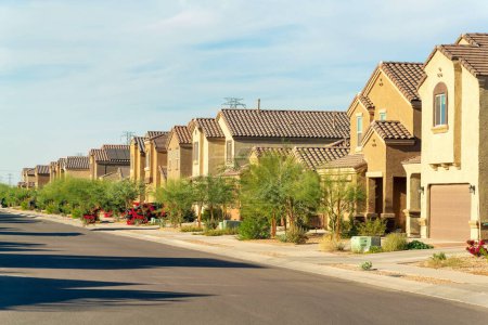 Row of suburban track home houses in a modern neighborhood with visible trees and clear blue sky. Stucco houses with warm desert colors white brown and beige in neighborhood.