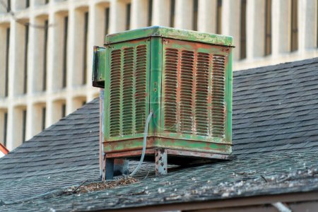 Téléchargez les photos : Swamp cooler air condition system on rooftop in modern city with green color and aged and weathered metal body. Gray roof tiles with white cement building background in afternoon shade. - en image libre de droit