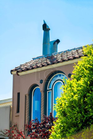 Photo for House facade or home large glass window with front yard tree and visible chimney pipes on orange stucco exterior. Late afternoon shade with bright tree and clear blue copy space sky. - Royalty Free Image