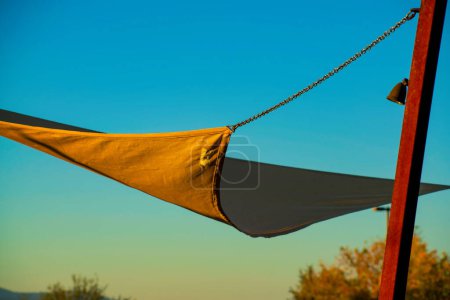 Overhang or tarp awning with cloth material hanging by metal beams and rope in late afternoon early morning sun. In urban or industrial area with hazy gradient orange and blue sky park or playground.