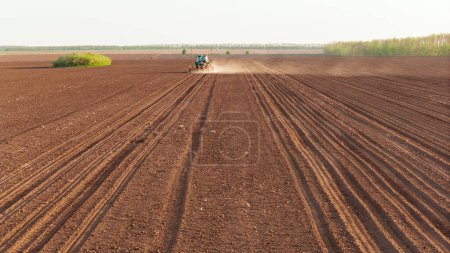 Aerial view of a tractor tilling the soil in a vast land lot, with a background of green grass, under a clear sky. Seeds to sow