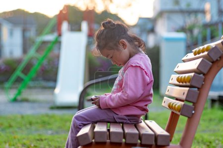 Foto de Un niño que usa o juega con un smartphone en el patio de recreo en lugar de jugar. Una niña pequeña viendo videos en un dispositivo inteligente. - Imagen libre de derechos