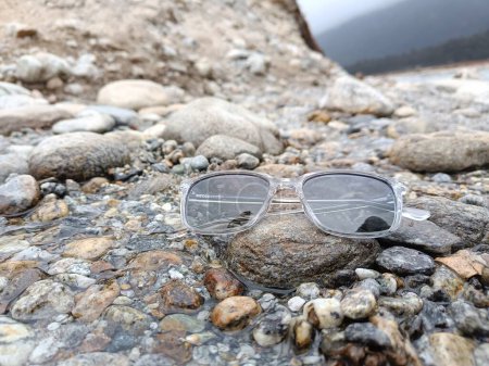Téléchargez les photos : A pair of modern, stylish, and trendy sunglasses or shades or sunnies placed on the top of a rock in a stream of water with beautiful natural background. The sunglasses have grey tinted lenses - en image libre de droit