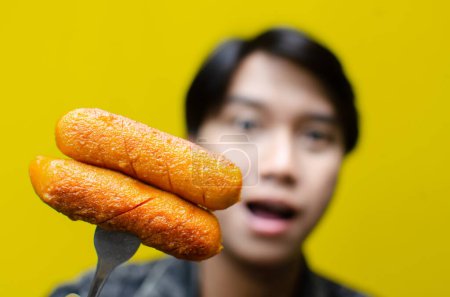 Téléchargez les photos : Foyer sélectif de l'homme asiatique mange et tient la saucisse sur une fourchette isolée sur fond jaune. concept de restauration rapide malsaine. - en image libre de droit