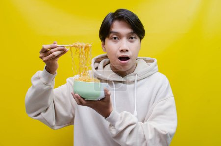 Photo for Closeup, an Asian man in a beige hoodie eats instant noodles using chopsticks and a bowl, isolated over a yellow background. - Royalty Free Image