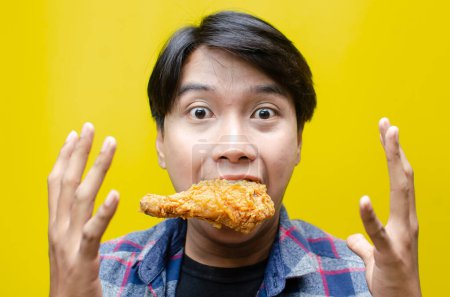 Téléchargez les photos : Extatique et expressif asiatique homme mange et mord poulet frit isolé sur fond jaune - en image libre de droit