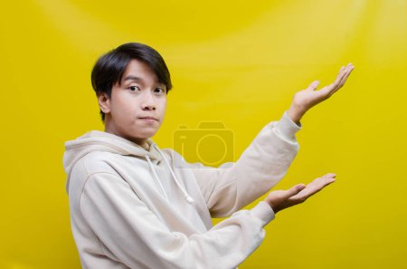 Foto de Vista lateral del hombre asiático en una sudadera con capucha beige señalando sus dedos y abriendo los brazos para promover y anunciar algo bueno. - Imagen libre de derechos