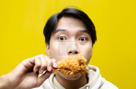 Foto de Extático y expresivo asiático hombre come y muerde frito pollo aislado en amarillo fondo - Imagen libre de derechos
