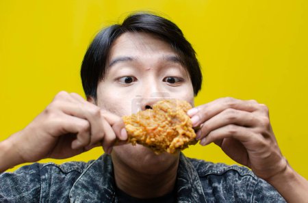 Photo for Ecstatic and expressive asian man eats and bites fried chicken isolated on yellow background - Royalty Free Image