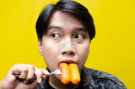 Foto de Extático y expresivo asiático hombre come y muerde salchicha usando tenedor aislado sobre amarillo fondo - Imagen libre de derechos