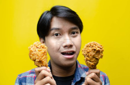 Foto de Feliz hombre asiático sorprendido sostiene pollo frito con expresión wow aislado sobre fondo amarillo. concepto de comida poco saludable. promoción de venta de alimentos - Imagen libre de derechos