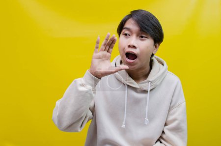 Téléchargez les photos : Asiatique homme portant un beige sweat à capuche crier et chuchoter pour annoncer de bonnes nouvelles. - en image libre de droit