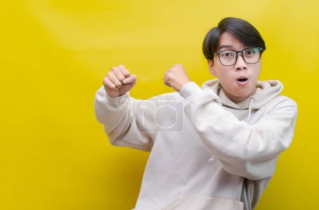 Téléchargez les photos : Homme asiatique en beige sweat à capuche dansant heureux et levant son poing célébrant la victoire sur fond jaune - en image libre de droit