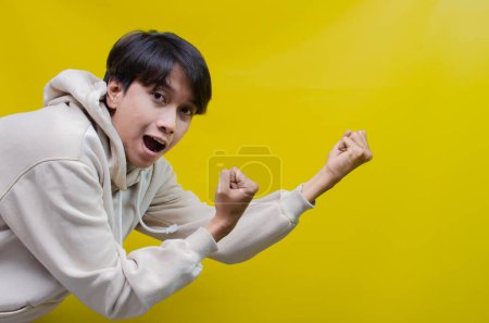 Foto de Asiático hombre en beige sudadera con capucha bailando felizmente y levantando su puño celebrando la victoria sobre fondo amarillo - Imagen libre de derechos