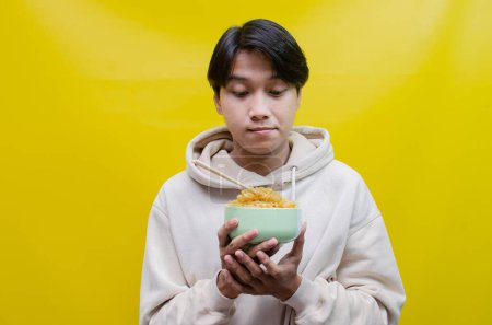 Photo for Closeup, an Asian man in a beige hoodie eats instant noodles using chopsticks and a bowl, isolated over a yellow background. - Royalty Free Image