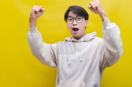 Foto de Asiático hombre en beige sudadera con capucha bailando felizmente y levantando su puño celebrando la victoria sobre fondo amarillo - Imagen libre de derechos