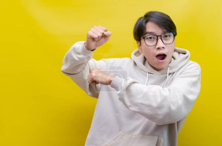 Téléchargez les photos : Homme asiatique en beige sweat à capuche dansant heureux et levant son poing célébrant la victoire sur fond jaune - en image libre de droit