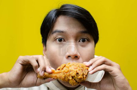 Photo for Ecstatic and expressive asian man eats and bites fried chicken isolated on yellow background - Royalty Free Image