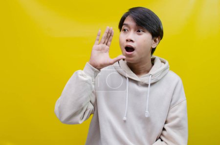 Photo for Asian man wearing a beige hoodie shouting and whispering to announce good news. - Royalty Free Image