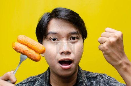 Téléchargez les photos : L'homme asiatique mange et tient des saucisses sur une fourchette isolée sur fond jaune. concept de restauration rapide malsaine. - en image libre de droit