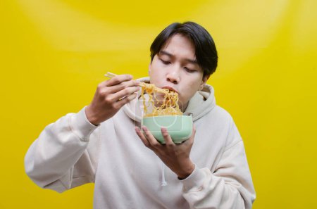Foto de Hombre asiático con la boca llena de fideos aislados sobre fondo amarillo. un hombre asiático en una sudadera con capucha beige come fideos instantáneos con hambre usando palillos y un tazón. - Imagen libre de derechos