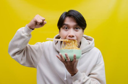 Photo for Asian man with mouth full of noodles isolated over yellow background. an Asian man in a beige hoodie eats instant noodles hungrily using chopsticks and a bowl. - Royalty Free Image