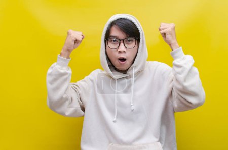 Photo for Asian man in beige hoodie dancing happily and raising his fist celebrating victory on yellow background - Royalty Free Image