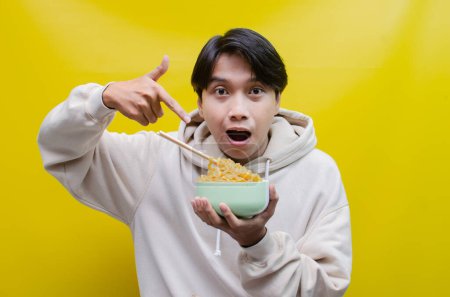 Téléchargez les photos : Gros plan, un asiatique en sweat à capuche beige mange des nouilles instantanées à l'aide de baguettes et d'un bol, isolé sur un fond jaune. - en image libre de droit