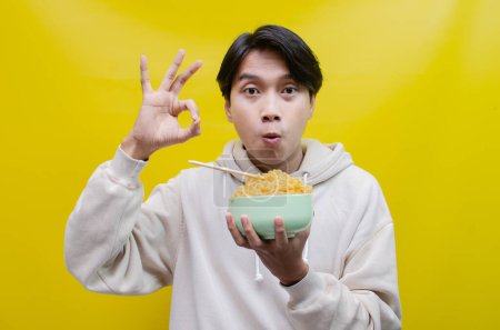 Téléchargez les photos : Gros plan, un asiatique en sweat à capuche beige mange des nouilles instantanées à l'aide de baguettes et d'un bol, isolé sur un fond jaune. - en image libre de droit