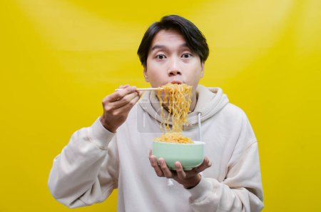 Téléchargez les photos : Homme asiatique avec la bouche pleine de nouilles isolées sur fond jaune. un homme asiatique dans un sweat à capuche beige mange des nouilles instantanées avec faim en utilisant des baguettes et un bol. - en image libre de droit